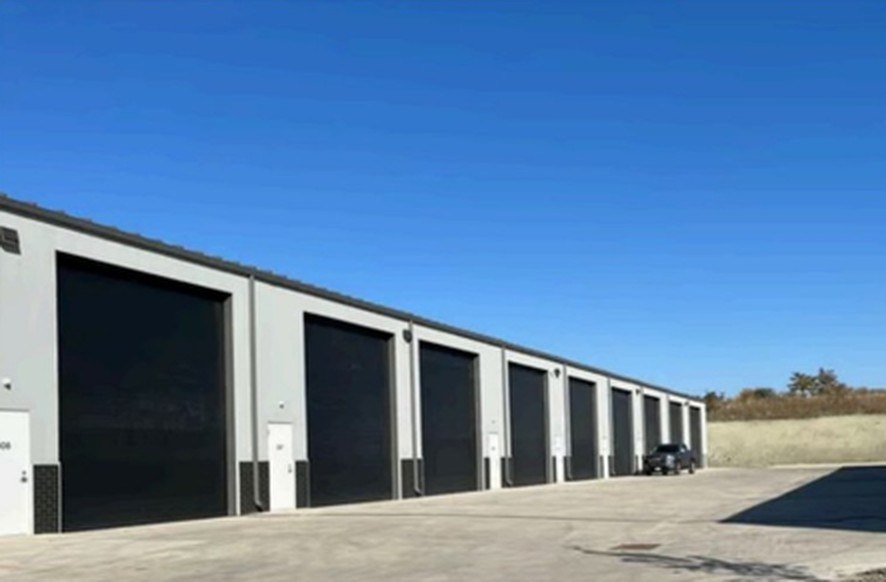 Row of gray metal contractor garages with tall black overhead doors along a wide concrete drive